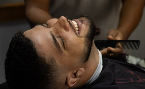 A client reclining in a barber chair while a barber combs his beard